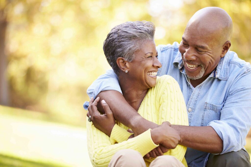 A senior man and woman sitting outdoors smile, with his arms around her shoulders.